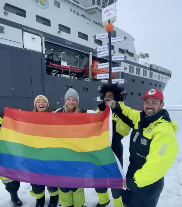 Five people standing on ice in front of a shop and wearing cold weather gear hold a rainbow pride flag in front of the North Pole marker.
