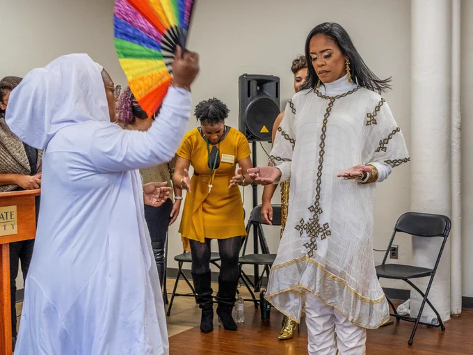 A medium shot of a woman with her head bowed, eyes closed, and hands outstretched wearing a white turtleneck, dress, and boots as she gets fanned by woman wearing an all-white hooded dress.