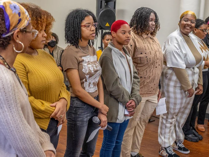 Six students stand side-by-side with their hands folded in front of them holding a piece of paper. One student in the center also holds a microphone at their side.