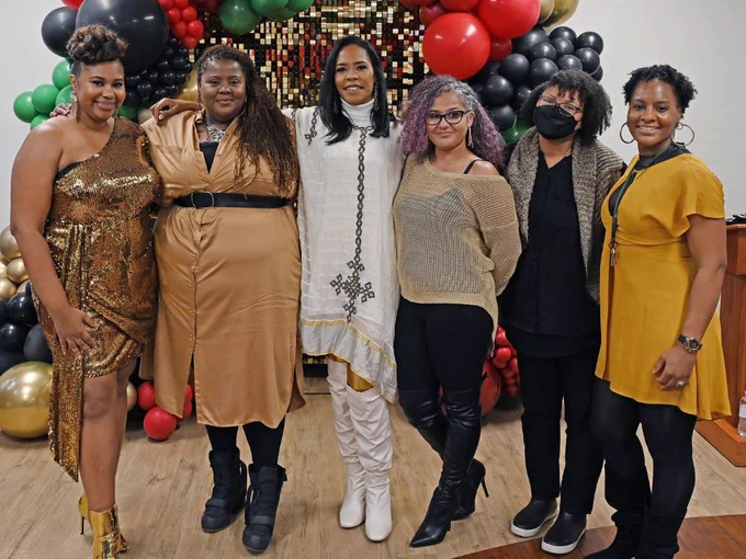 A full shot of six smiling women standing side-by-side in front of a red, green, gold, and black balloon installation.