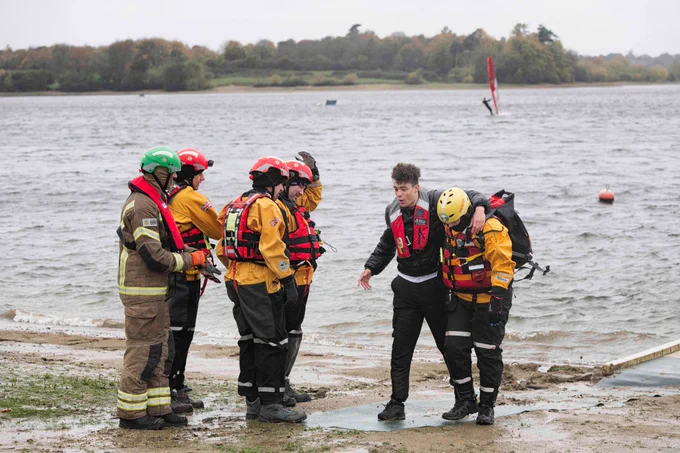 People from Suffolk Fire and Rescue Service and Suffolk Lowland Search and Rescue help a student to shore following a water rescue at Alton Water. A large body of water, trees and a windsurfer can be seen in the background.