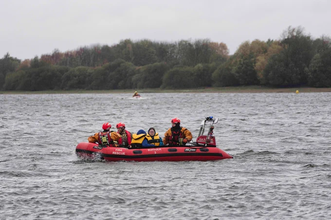 A boat containing Suffolk Fire and Rescue Service and Suffolk Lowland Search and Rescue staff transports rescued college students on Alton Water. Trees and a further boat can be seen in the background. 