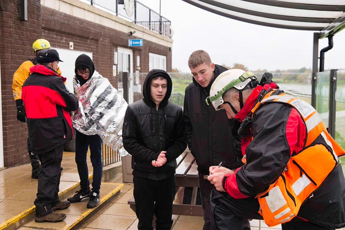 Staff from Suffolk Fire and Rescue Service and Suffolk Lowland Search and Rescue help three students who have been rescued from Alton Water. They are stood huddles under what appears to be a gazebo. One of the students is wearing a foil blanked for warmth.