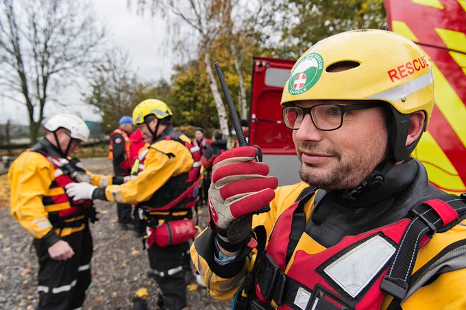 A man wearing a helmet, glasses, a wet suit, life jacket and gloves looks pensive while holding a walkie talkie. Trees and colleagues from Suffolk Fire and Rescue Service and Suffolk Lowland Search and Rescue can be seen in the background.