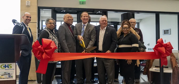 A smiling group of people stand behind a large red ribbon stretched between two bows. A man holding a giant pair of yellow scissors prepares to cut the ribbon.