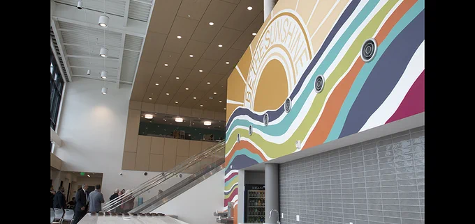 A mural is painted with the text "be the sunshine" over a sun and above rows of brightly colored waves. It is in the foreground of the brightly lit interior of a building with people gathered in the background.