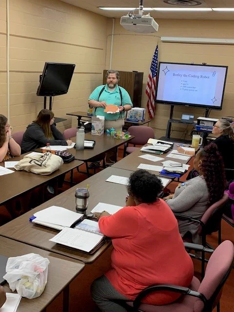 A white man with a beard and a teal shirt stands in front of a PowerPoint presentation. Two rows of tables are in front of the presentation with various librarians sitting behind the tables.