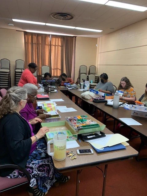 Two rows of tables sit in the middle of a conference room. Various librarians sit behind the table, looking at various notebooks and binders.