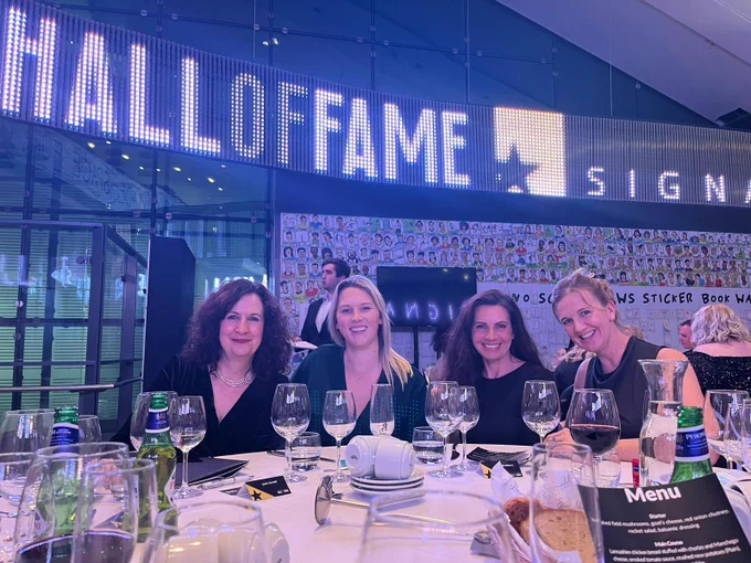 Four ladies smiling whilst sat at a table with wine glasses, beer bottles, ad bread baskets. Behind them is a large digital display with the text ‘Hall of fame, Signature’.