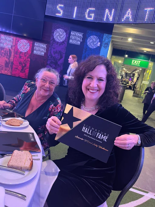 Two women smiling whilst sitting at a dinner table. One woman is proudly holding up a large card branded with the Hall of Fame colours and text. The card states the shortlisted individual, which for this card is Lipspeaker UK and Lesley Weatherson.