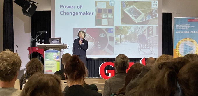 Julia Kleeberger auf der Bühne mit Mikrofon, im Hintergrund ihre Präsentationsfolie mit dem Titel "Power of Change Maker". Im Vordergrund zuhörende Teilnehmende.