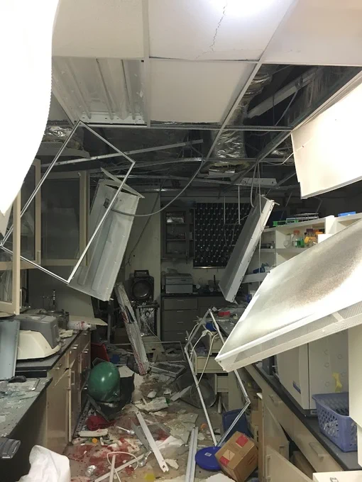 A laboratory in ruins:  explosion has damaged ceiling, and panels hang chaotically from their frame.  Boxes, debris and detritus are strewn across the floor.