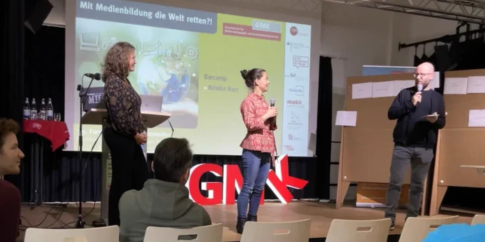 3 Menschen auf der Bühne bei der Anmoderation. Im Hintergrund die Buchstaben G, M und K und eine Präsentationsfolie.
