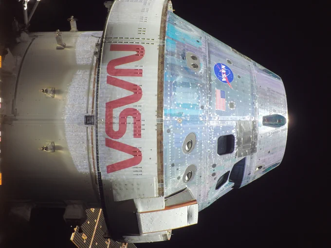 In this Orion "selfie," part the Orion spacecraft is viewed from the side against the blackness of space. The outside of the spacecraft is white silver and shaped like a stout cone with a cylinder base. Windows are visible on the side of the spacecraft, as well as an American flag, a NASA meatball logo, and the word "NASA" in red letters. Part of a gold solar array can be glimpsed behind the spacecraft.