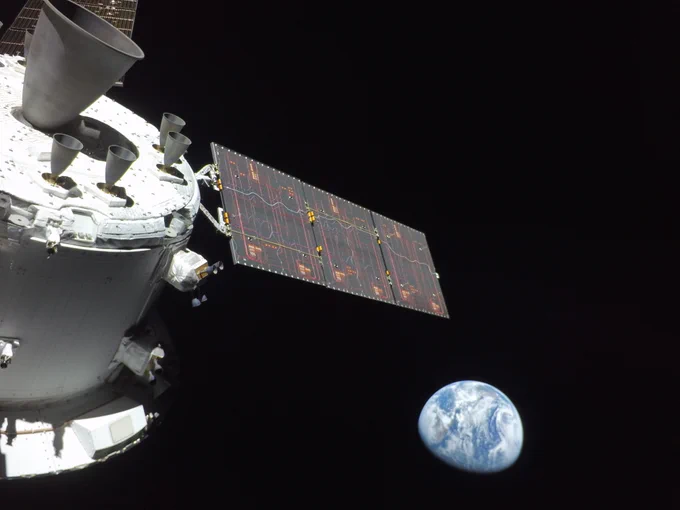 A color view of Earth as seen from Orion. The top half of Earth is illuminated, meaning the orientation of the image is tilted. The spacecraft's solar panel extends into the image along with some of its thrusters. 
