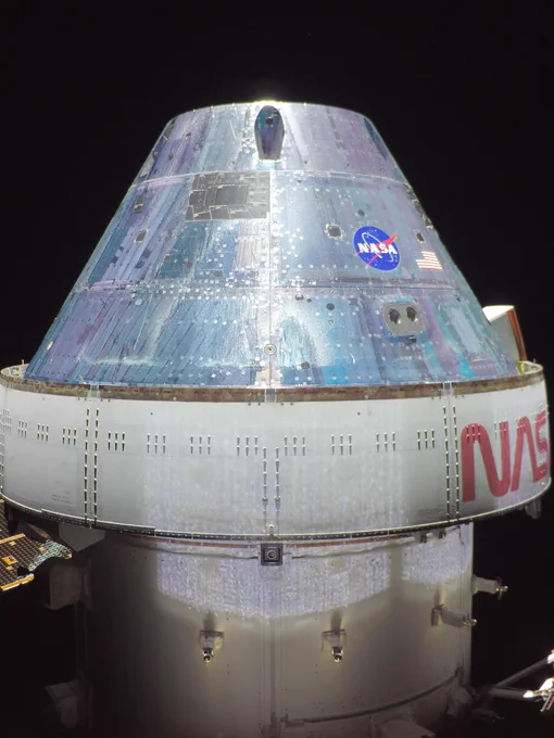 In this Orion "selfie," part the Orion spacecraft is viewed from the side against the blackness of space. The outside of the spacecraft is white silver and shaped like a stout cone with a cylinder base. 