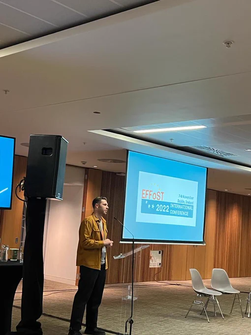 The picture shows Mark Al-Shemmeri addressing the audience during his presentation. The logo of the conference is displayed in the screen at his side.