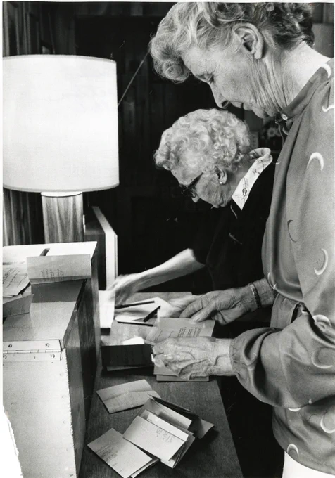 Two women are standing at a table, counting a stack of paper ballots.