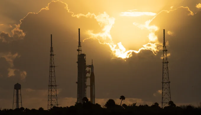 The Space Launch System rocket and Orion spacecraft are silhouetted against a gold sky. The white light of the Sun is breaking through the clouds in the distance. To the rocket's left, sit a water tower and lightning tower. To the rocket's right, sits another lightning tower. 