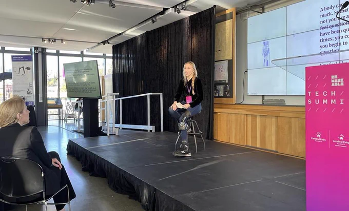 a photo of Katy, a white woman with blonde hair and dark clothes on and she’s sitting on a stool placed on a small platform stage. She wears an assistive device on her left leg to support her mobility with cerebral palsy. The podium for the Remarkable Tech Summit appears to the right and a screen is behind her featuring a quote she used in her speech.