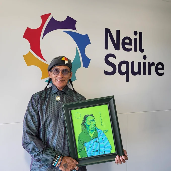 Ron holding one of his pieces of art, smiling in front of the Neil Squire logo
