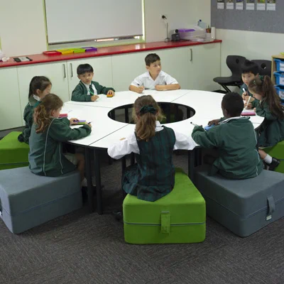 8 students sitting around 4 quarter circle tables, arranged in a circle. The table is arranged in a circle and students are sitting on grey and green wedge-shaped soft ottomans. The students' uniform is green, also.