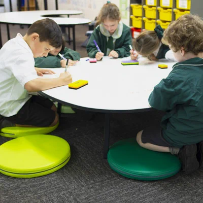 5 young students kneeling on flat cushions at a low table. The table is writable and each student has a texta and eraser ready. The students are looking down and they are doing individual writing work