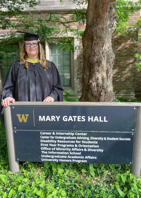 Jamie Poirier stands in her graduation cap and gown outside Mary Gates Hall. She is a a white woman with glasses and blonde hair.