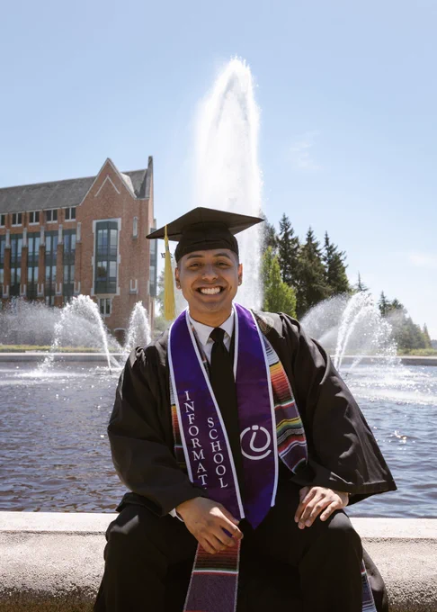 Diego smiles in his grad cap and gown in front of Drumheller Fountain.