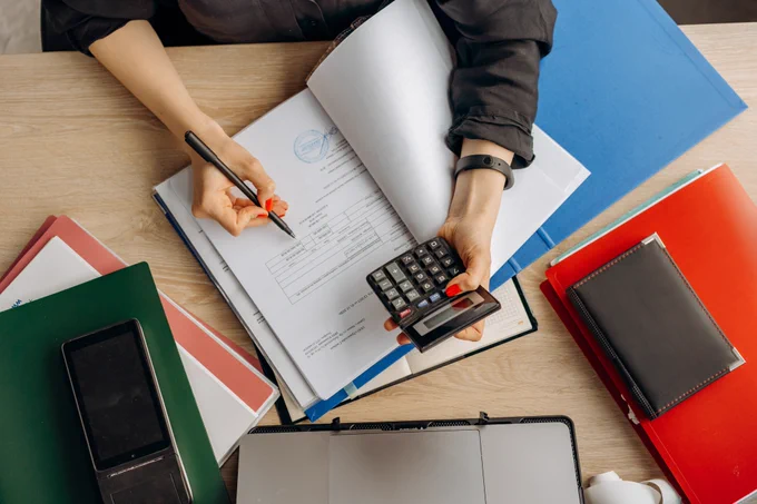 A close up photo of a woman working at their desk with a calculator in their left hand and a pen in their right. They are working through some paper documents.