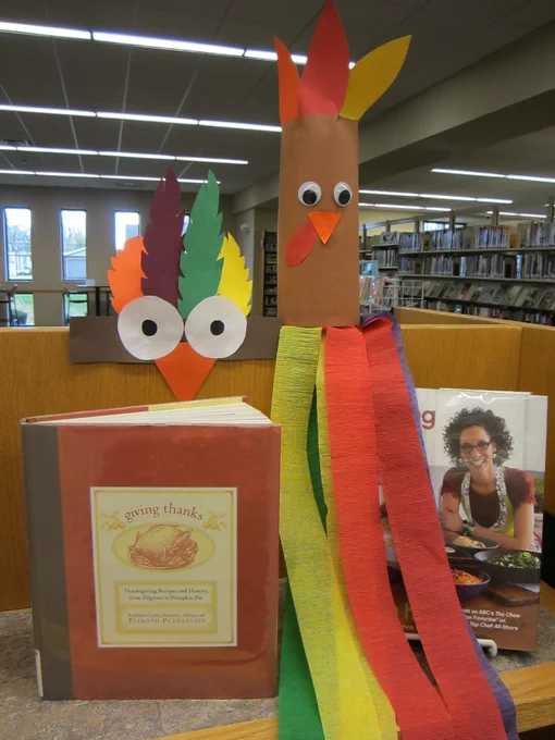 A brown paper cylinder with googly eyes and an orange triangle beak. It has feathers on top and long colorful streamers at the bottom. It's positioned with a book called Giving Thanks