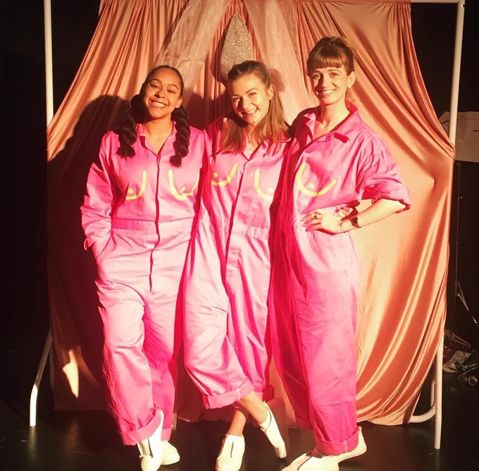 Three women in pink boiler suits with yellow boobs painted on, standing infront of a curtain shaped like a vulva 