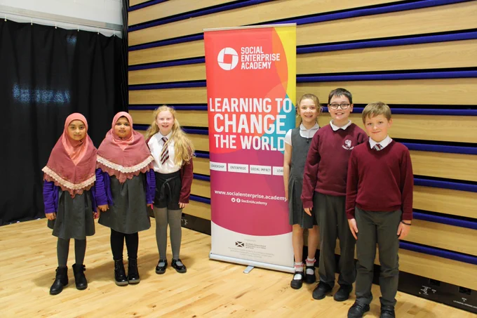 Three primary school pupils stand either side of a Social Enterprise Academy banner that reads 'Learning to change the world'. They are on a light wooden floor with a striped wooden wall behind them. 