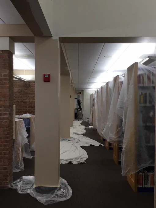 Plastic tarps covering non-fiction book shelves with a painter up on a ladder in the far back.
