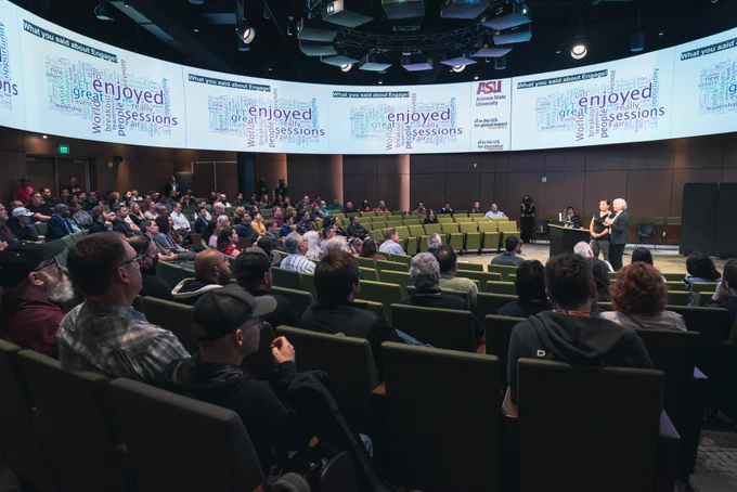 View of the room at Tempe campus, with speakers at front and screens around the top.