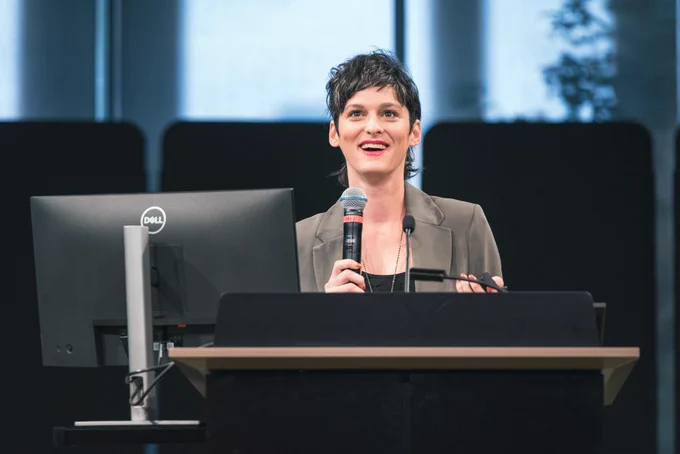 Woman in grey suit jacket on stage presenting with mic in hand