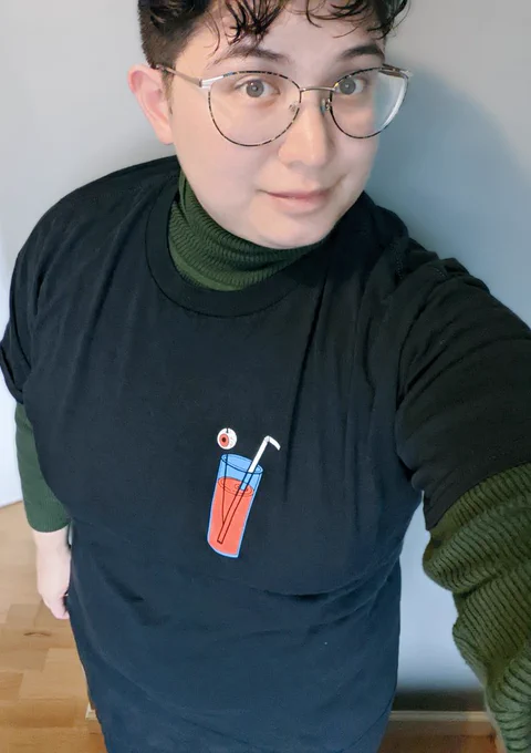 Élaina takes a selfie wearing a black t-shirt wit a cartoon glass of blood with an eyeball and straw on the left breast.