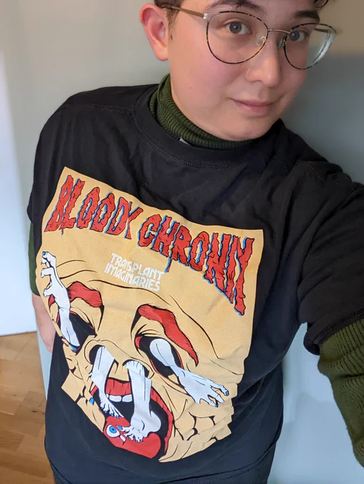 Élaina takes a selfie wearing a black t-shirt that has a graphic of a white body coming out of a grotesque face holes. Aboverare the words "Bloody Chronic" in red bloody letterring.