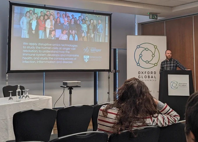 Picture of Dave standing on the right hand side of a conference room infront of an 'Oxford Global' banner. His arm out as he speaks over a presentation slide showing a photograph of the Haniffa lab and the lab aims