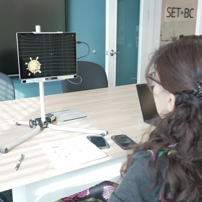 SET-BC educator Kamelia sitting by a desk in front of  a tablet mounted to a desktop mount, with the PCeye 5 tracker camera attached. 