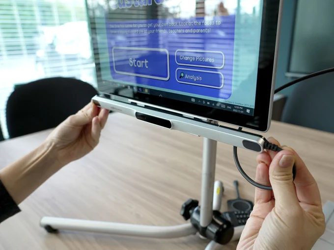 Two hands hold the PCEye 5 tracker camera, a long finger-thin piece of technology, in front of a tablet screen on a mount. The eye tracking software can be seen on the screen..