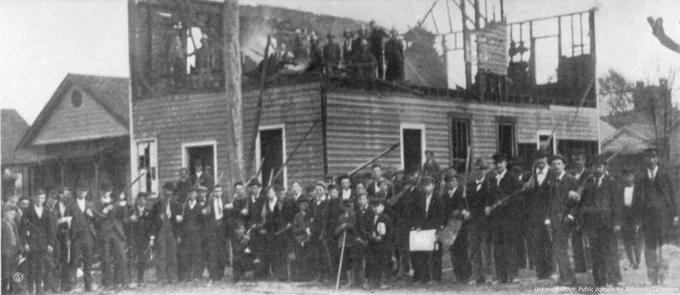 A black and white photo of the wrecked "Record" building behind a large group of vigilantes from the 1898 Wilmington Massacre.