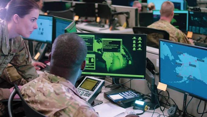 Two Soldiers look at a pair on monitors with maps on them. They are in a control with other soldiers looking at computers. (Photo Credit: Maj. Christopher Vasquez, U.S. Air Force)