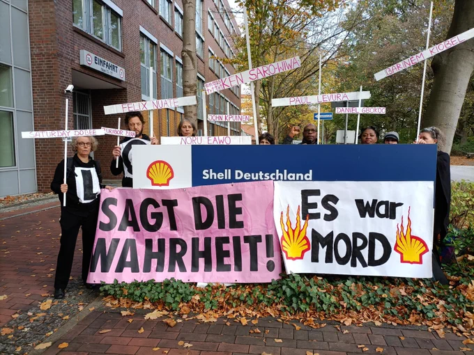 8 Menschen stehen hinter einer Anzeigetafel auf der das Shell-Logo und "Shell Deutschland" steht. Sie halten zwei Banner: "Sagt die Wahrheit!" und "Es war MORD". Auf dem zweiten Banner ist das Shell-Logo in Flammen zu sehen. Einige Menschen halten weiße Kreuze auf denen verschiedene Namen stehen "Ken Saro-Wiwa", "Saturday Dobee", "Nordu Eawo", "Daniel Gbooko", "Paul Levera", "Felix Nuate", "Baribor Bera", "Barinem Kiobel" und "John Kpuine".