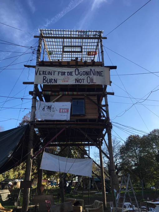 You can see a large wooden tower in front of a blue sky and trees in the background. On the tower hangs, among other things, a banner with the text "United for the OgoniNine - Burn Shell not Oil (Vereint für die OgoniNine - Brun Shell not Oil)" with the Shell logo in flames above Shell.