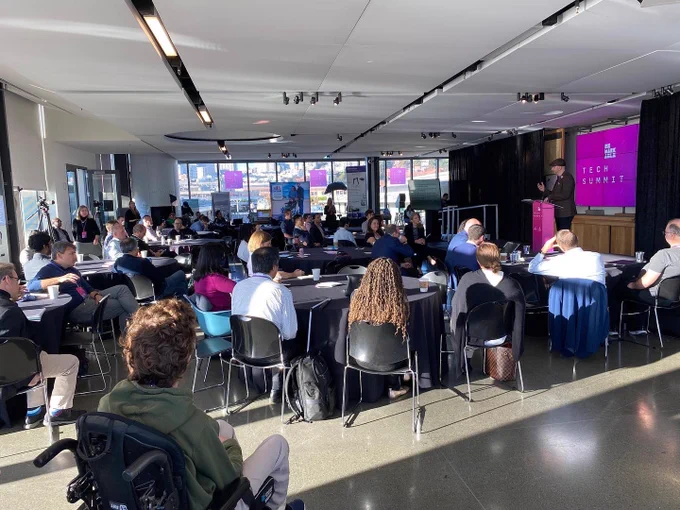 a photo of the Fisher Bay Observatory at the Exploratorium in San Francisco with attendees from the Remarkable Tech Summit. Pete, the Founder of Remarkable stands on a platform stage at a podium speaking to the audience. There’s lots of round tables and light coming in from big windows at all angles.