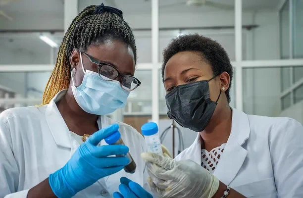 icipe-supported PhD students Linda Bih Numfor of Cameroon and Ruth Lorivi Moirana of Tanzania, both wearing white lab coats, face masks, and gloves, hold up lab tubes containing liquids. (Photo courtesy of icipe)