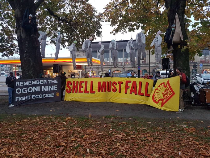 Im Hintergrund ist eine Shell-Tankstelle zu sehen. Zwischen zwei großen Bäumen ist ein Seil gehängt, an dem 9 weiße Overalls hängen, die wie erhängte Menschen aussehen. Auf dem Boden stehen einige Menschen mit Bannern: "Remember the Ogoni Nine - Fight Ecocide" mit brennendem Shell-Logo in einer Ecke, "Shell must fall" und "Shell must fall" in einem großen Shell-Logo.
