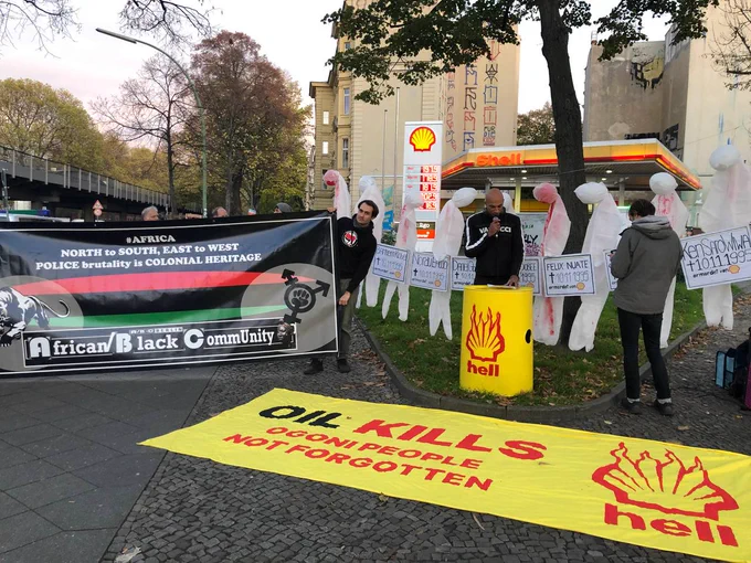Im Hintergrund ist eine Shell-Tankstelle zu sehen. Links halten Menschen ein Banner mit dem Text "#Africa - Nort to Sotuh, East to West - Police brutality is Colonial Heritage - African/Black Community". Auf dem Banner ist auch noch ein Panther, eine rot-schwarz-grün gestreifte Flagge und ein Transgender-Zeichen mit einer Faust im Kreis zu sehen. Rechts ist ein Seil gespannt, an dem 9 blutige weiße Overalls hängen, die wie erhängte Menschen aussehen. Davor steht eine Person an einem Ölfass und hält eine Rede. Auf dem Ölfass ist ein brennendes Shell-Logo zu sehen unter dem "hell" steht. Davor liegt auf dem Boden ein Banner mit einem brennenden Shell-Logo unter dem "hell" steht und dem Text "Oil Kills - Ogonia People Not Forgotten".