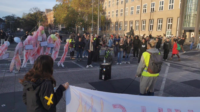 Im Hintergrund ist ein größeren Gebäude zu sehen. Davor stehen einige Dutzend Menschen um eine andere Person mit Mikro in der Hand herrum. Links halten ein paar Menschen ein Seil gespannt, an dem 9 blutige weiße Overalls hängen, die wie erhängte Menschen aussehen. Ganz vorne im Bild ist eine Person von hinten zu sehen, die ein Banner hält.